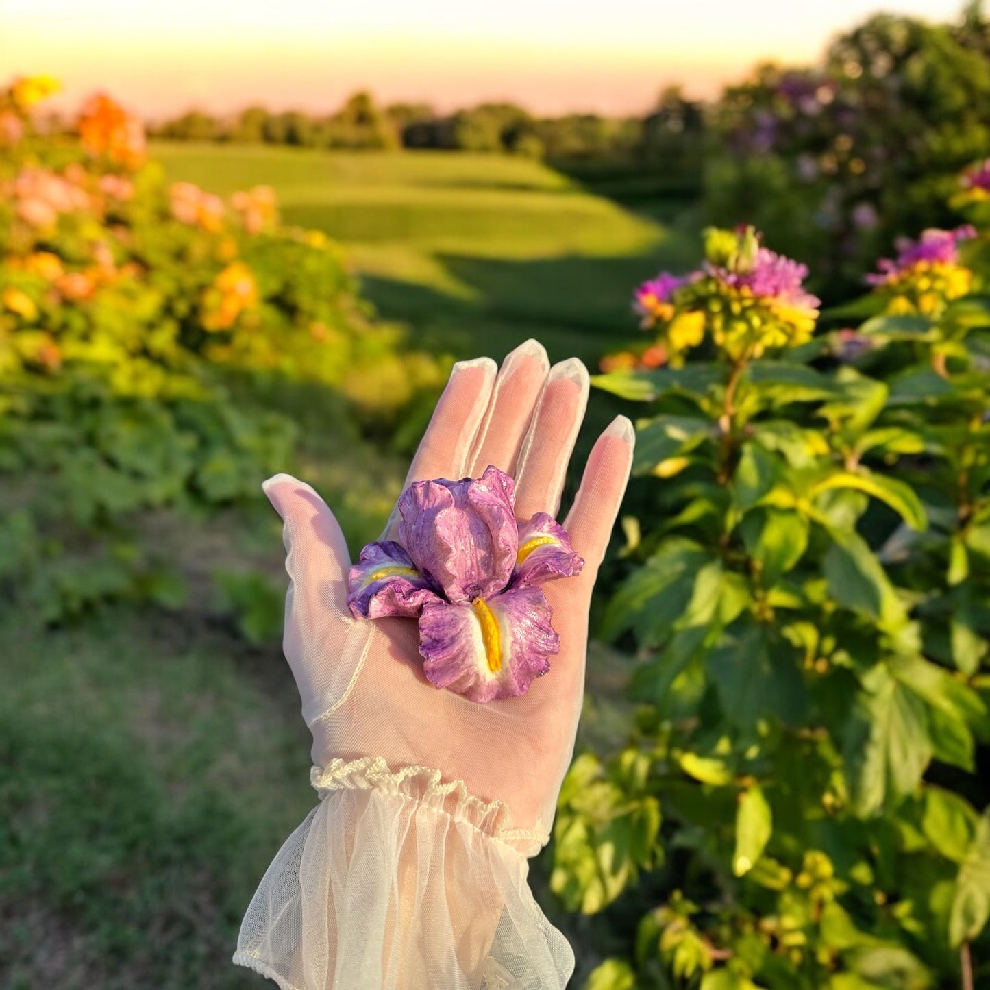 Hand holding a purple marshmallow flower resembling an iris, outdoors in a garden with greenery and flowers in the background.