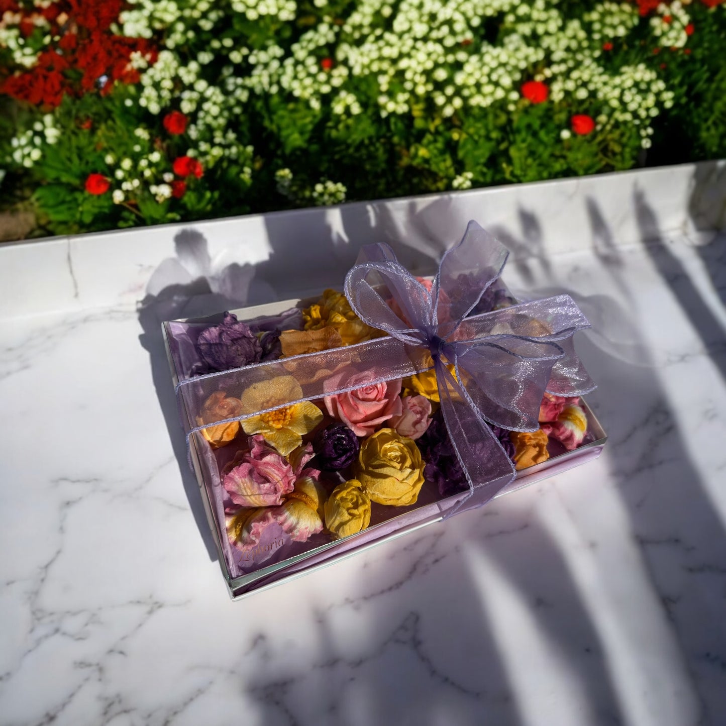 Clear gift box filled with colorful marshmallow flowers tied with a ribbon, placed on a marble surface outdoors with flowers in the background.