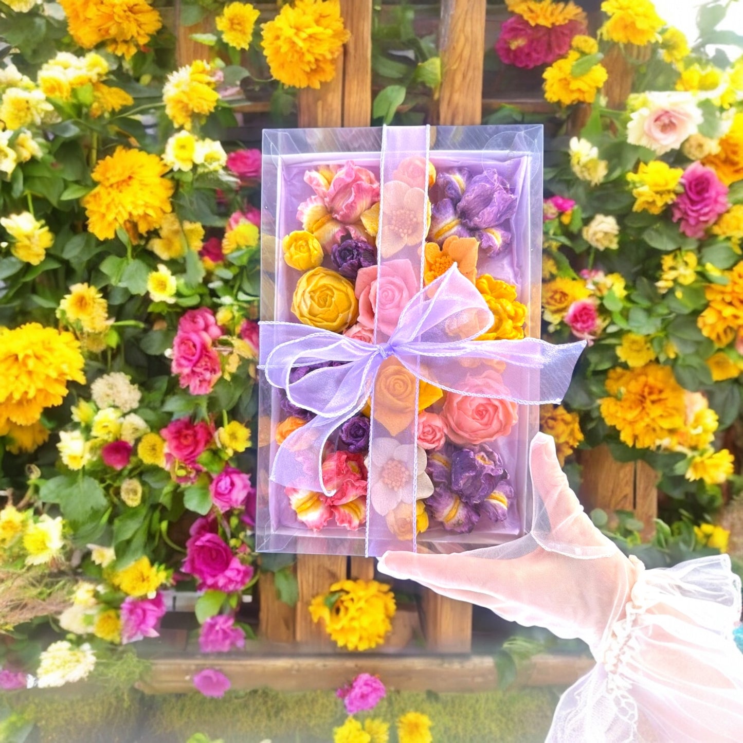 Hand holding a clear gift box filled with colorful marshmallow flowers, tied with a light purple translucent ribbon, against a background of blooming garden flowers.